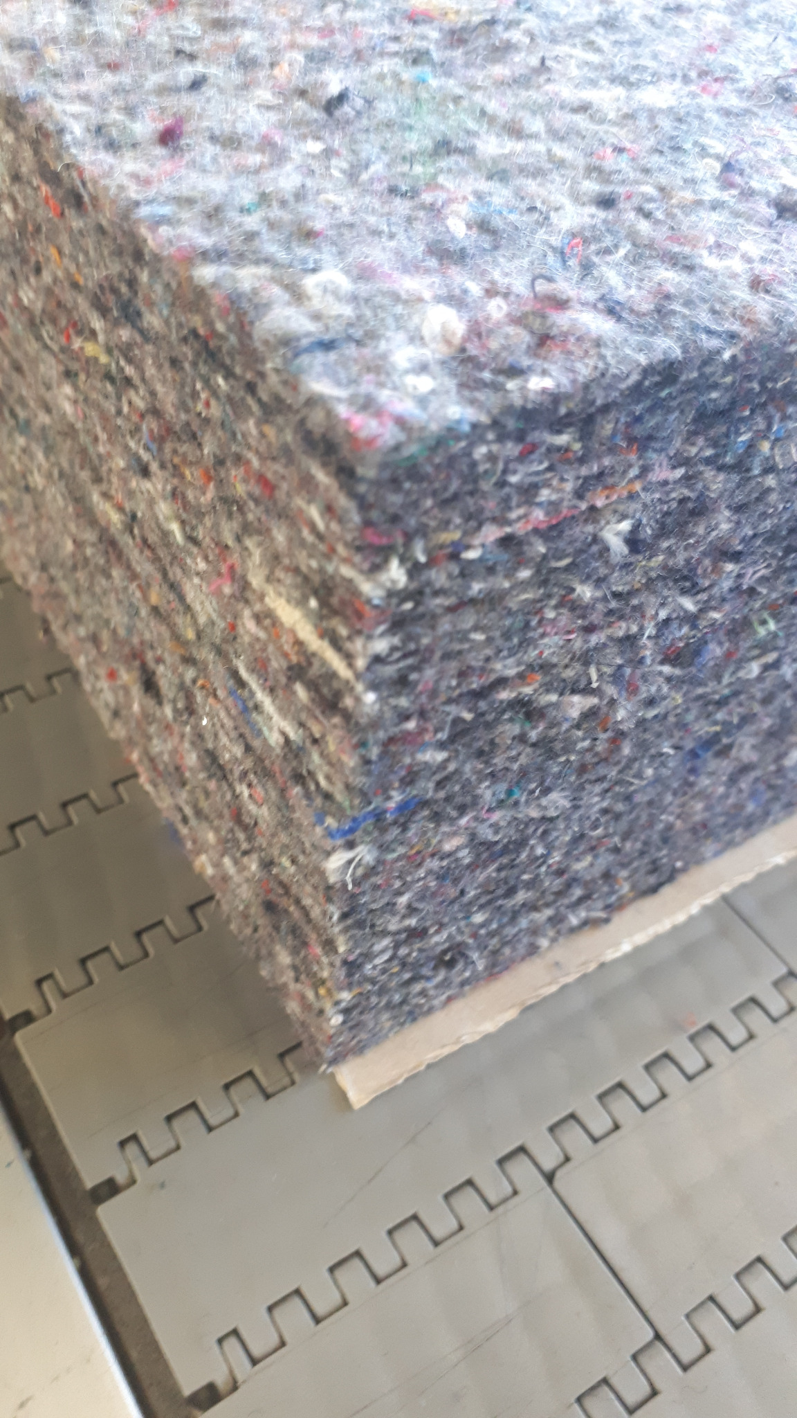 Blok szarego filcu technicznego z kolorowymi włóknami, ułożony na palecie transportowej z tworzywa sztucznego.