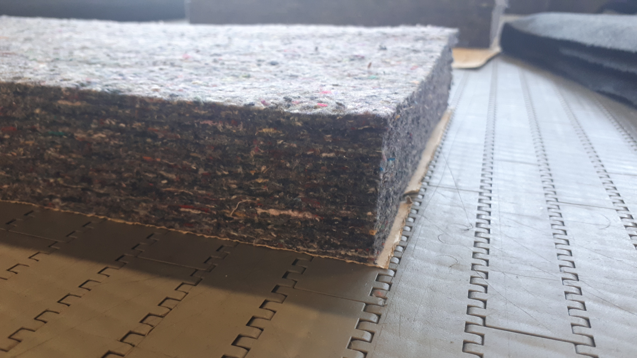 Wielowarstwowa płyta filcowa o nieregularnej strukturze i widocznych włóknach w różnych kolorach, leżąca na szarej, modułowej podłodze przemysłowej.