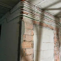 Równoległe wiązki białych kabli elektrycznych z zielonymi przewodami, mocowane do surowego, betonowego sufitu i ceglanych ścian w trakcie remontu.
