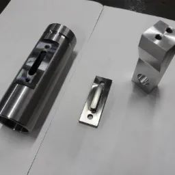 Precyzyjnie obrobione, metalowe elementy: cylinder z otworem, prostokątna płytka z podłużnym wgłębieniem i prostopadłościan z otworami, ułożone na białym papierze.
