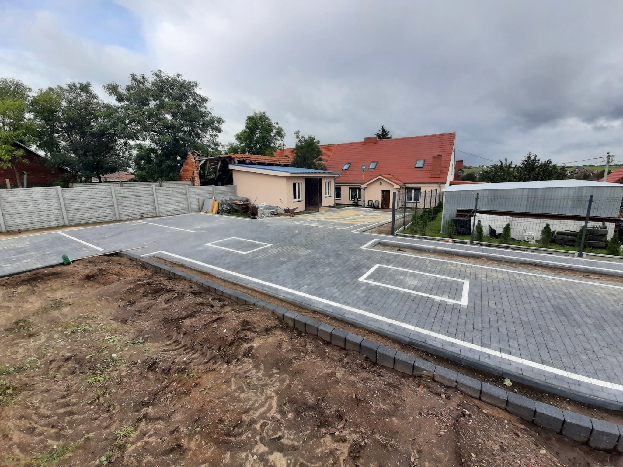 Nowo wyłożony, szary plac z kostki brukowej z białymi obramowaniami, widok z góry, obrzeża z ciemnej kostki, w tle budynki mieszkalne i pochmurne niebo.