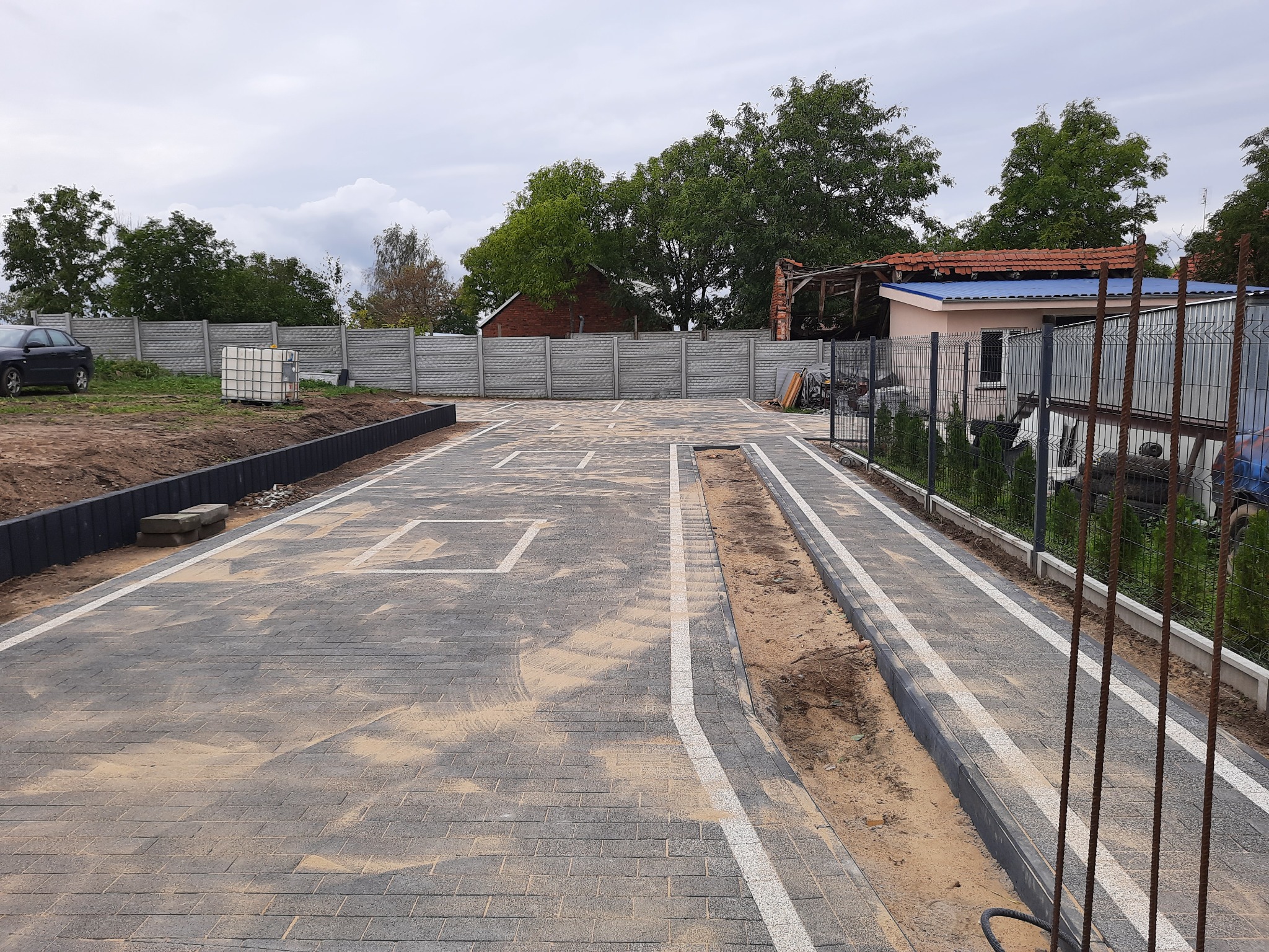 Wykończona nawierzchnia z szarej kostki brukowej z wyznaczonymi białymi liniami, tworzącymi miejsca parkingowe, obrzeża z kostki granitowej, widok na posesję z ogrodzeniem betonowym i metalowym...