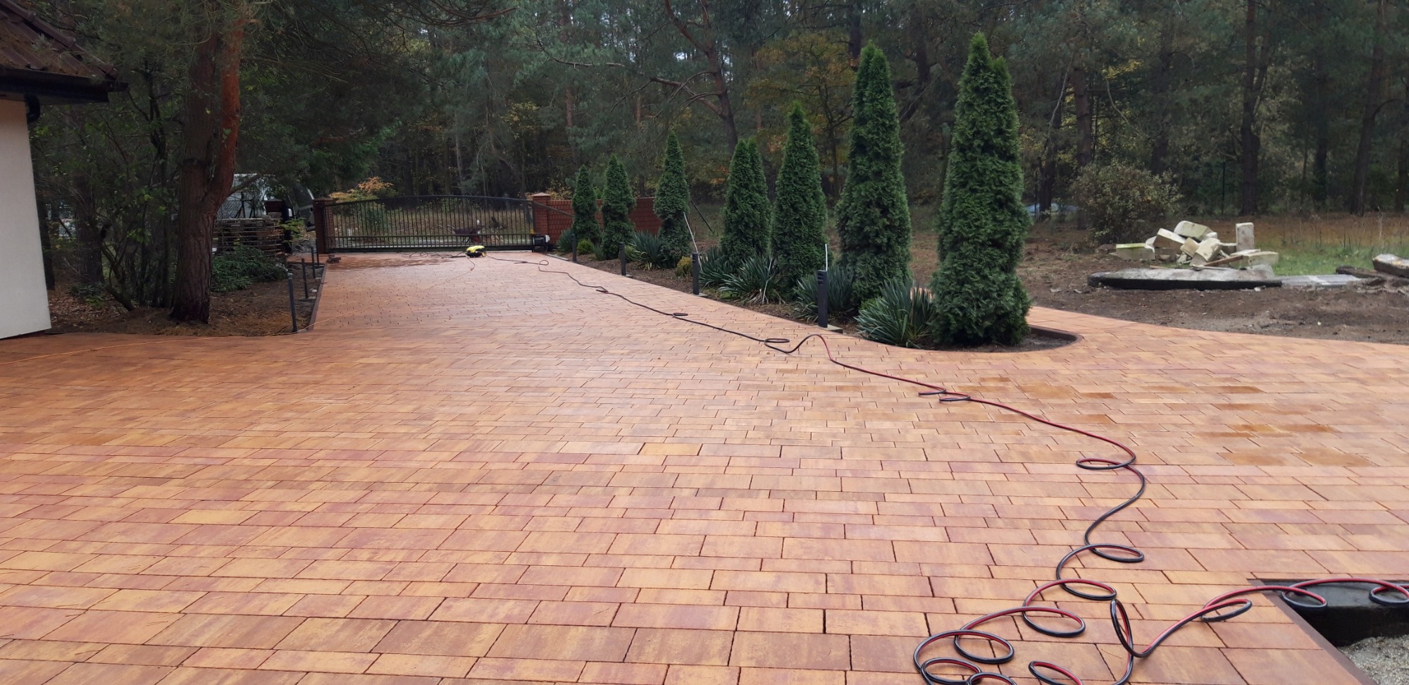 Nowo ułożona, pomarańczowo-brązowa kostka na podjeździe w Pobiedziskach, zakończona rabatą z tujami i ozdobnymi trawami, widoczny zwinięty wąż ogrodowy.