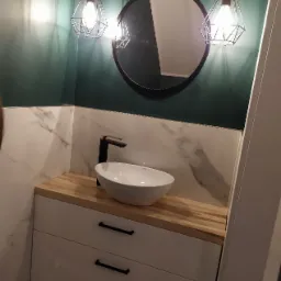 Nowoczesna łazienka: biała umywalka nablatowa z czarną armaturą na szafce z drewnianym blatem, marmurowe płytki na ścianie, okrągłe lustro i dwie lampy wiszące w stylu industrialnym na tle...