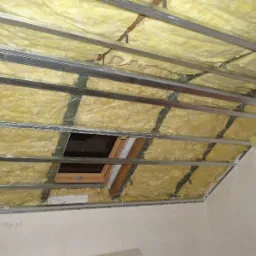 Poddasze w trakcie ocieplania wełną mineralną, widoczna konstrukcja metalowa i okno dachowe, poziomica oparta o ścianę.