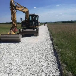 Żółta koparka CAT na drodze wysypanej białym kruszywem, widoczny wał drogowy w oddali, zielone pola po bokach.