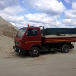 Czerwony samochód ciężarowy MAN z pustą skrzynią ładunkową stoi na piaszczystym terenie, obok góry piasku i lasu na horyzoncie, pod błękitnym niebem z chmurami.