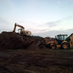 Żółta koparka CAT na szczycie pryzmy ziemi, obok koparko-ładowarka w trakcie prac ziemnych na polu w pochmurny dzień.