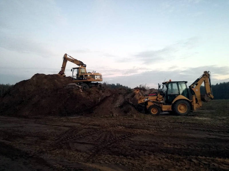 Żółta koparka CAT na szczycie pryzmy ziemi, obok koparko-ładowarka w trakcie prac ziemnych na polu w pochmurny dzień.