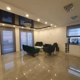 Elegancki salon z szarymi roletami rzymskimi na oknach, zielonymi sofami i błyszczącą podłogą, oświetlony przez nowoczesne lampy sufitowe i żyrandol.