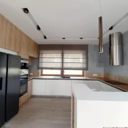 Nowoczesna kuchnia z szarą ścianą, białymi szafkami, drewnianym blatem i oknem przesłoniętym beżową tkaniną zwijaną w poziome pasy.