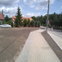 Nowo ułożona ścieżka z jasnoszarej kostki brukowej prowadząca wzdłuż drogi asfaltowej, z przygotowanym pasem ziemi pośrodku, w tle domy jednorodzinne i drzewa.