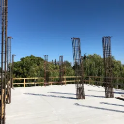 Zbrojenie pionowe wystające z betonowej płyty stropowej na budowie, w tle zielone drzewa i robotnicy w kaskach.