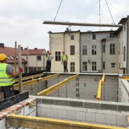 Budowa domu z białych bloczk&oacute;w: robotnicy w ż&oacute;łtych kaskach i kamizelkach odblaskowych nadzorują podnoszenie prefabrykowanego elementu stropu dźwigiem, widoczne drewniane podpory i szalunki, w tle...