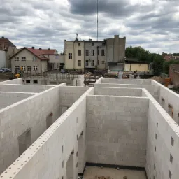 Wewnętrzne ściany domu w trakcie budowy, wzniesione z białych bloczków betonu komórkowego, widoczne otwory okienne i drzwiowe, w tle zabudowa miejska i pochmurne niebo.