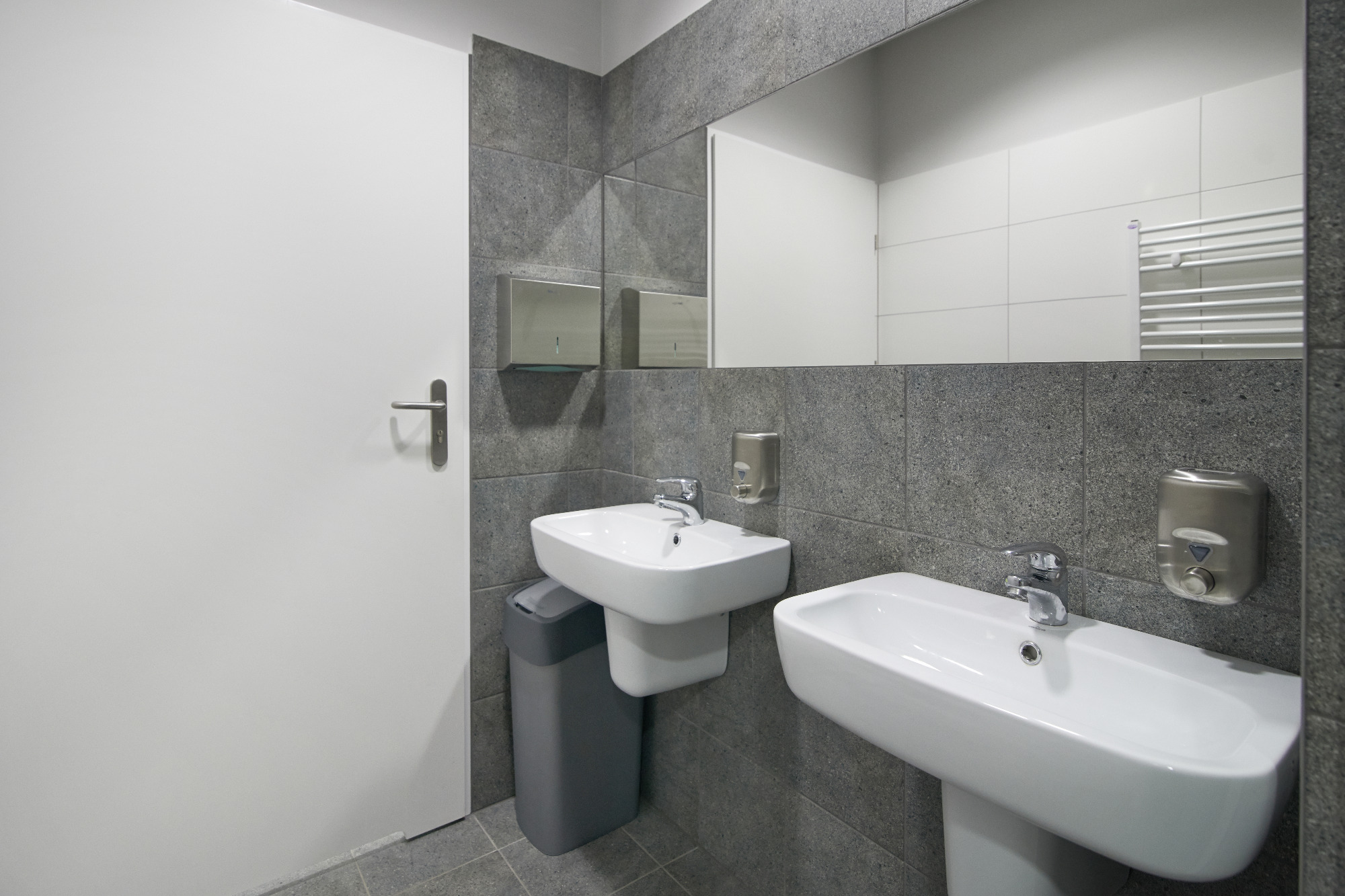 Nowoczesna łazienka publiczna z dwiema białymi umywalkami, szarą mozaikową ścianą i lustrem, w którym odbija się grzejnik drabinkowy i białe płytki.