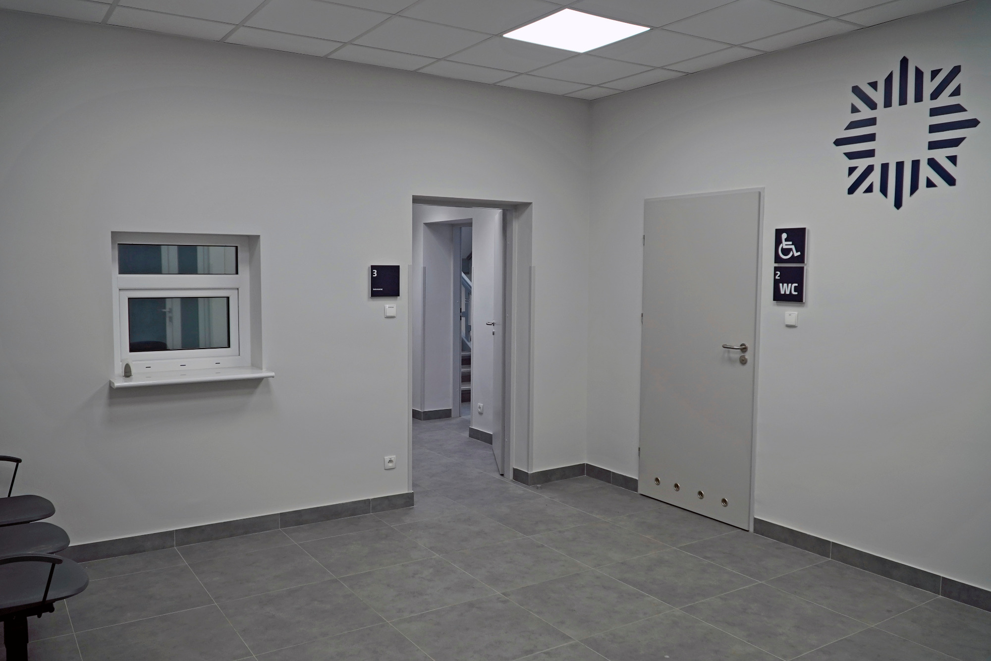 Wnętrze biurowe z szarą podłogą, białymi ścianami, oknem podawczym, drzwiami z oznaczeniem WC dla niepełnosprawnych i dekoracyjnym znakiem graficznym na ścianie.