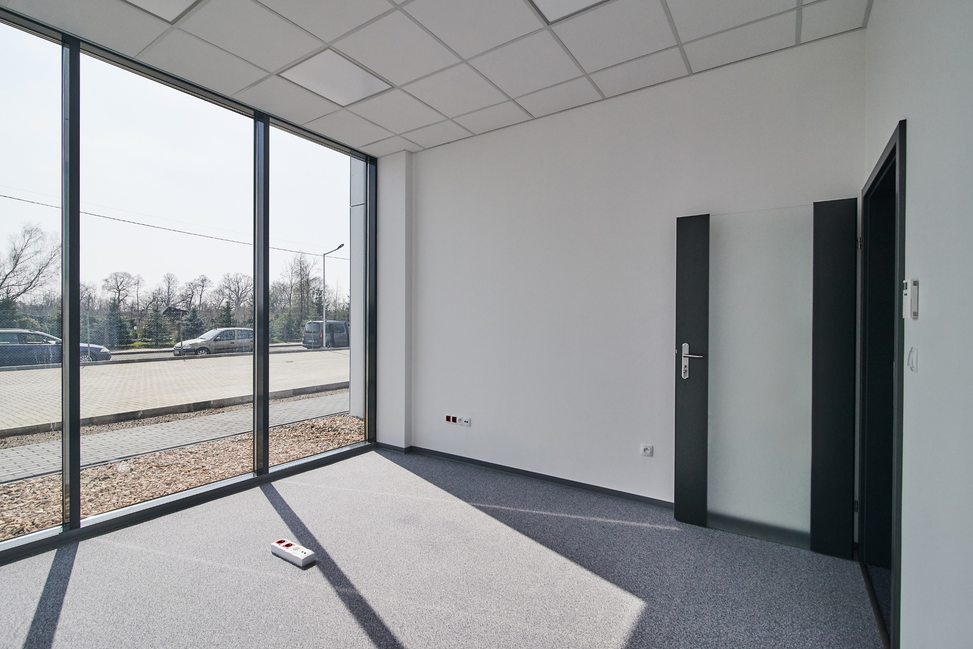 Wnętrze biura po remoncie z szarą wykładziną, dużym oknem z widokiem na parking i częściowo otwartymi drzwiami z matowego szkła.