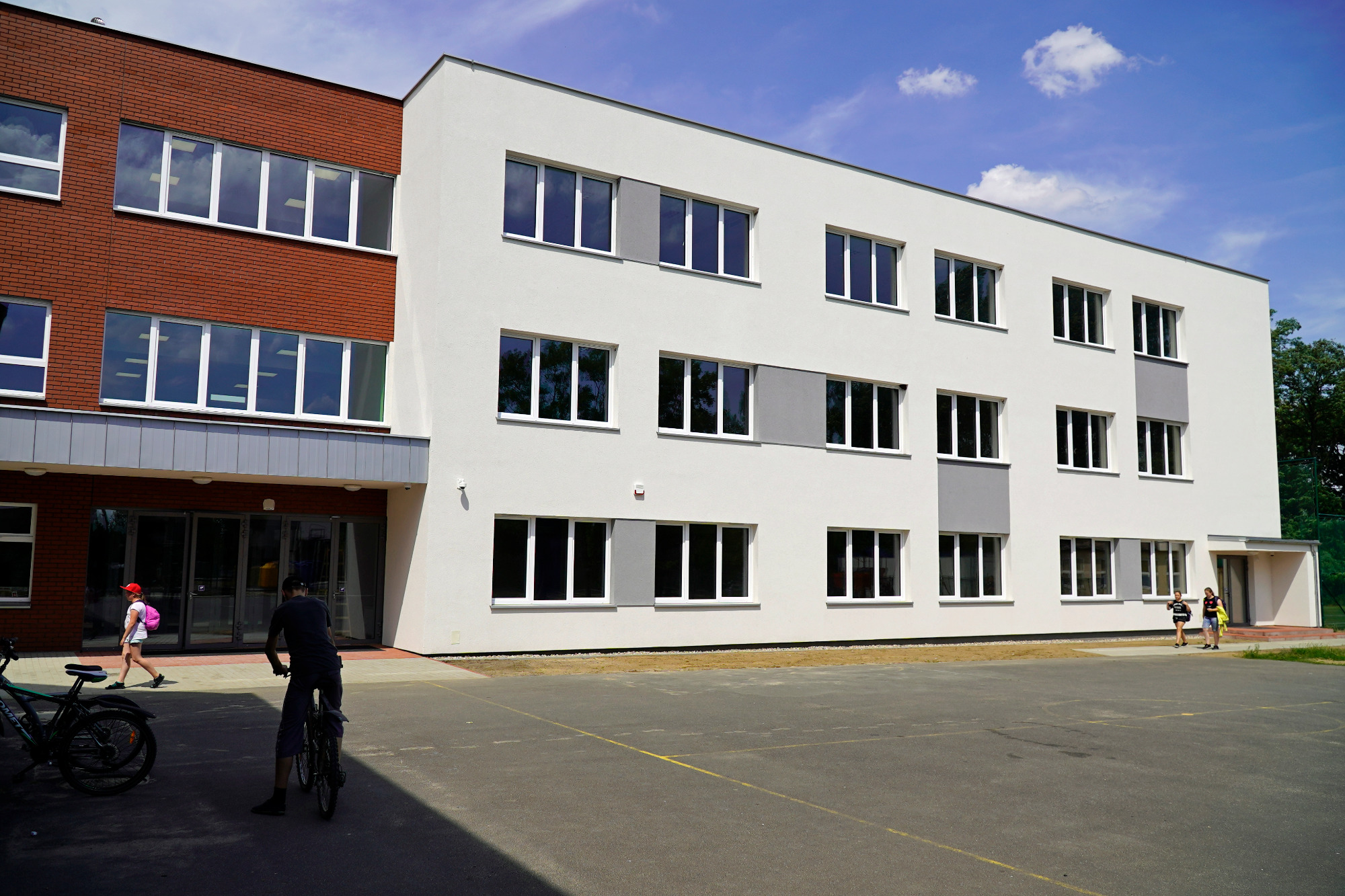 Nowoczesny budynek szkoły podstawowej z elewacją w kolorze białym i ceglastym, widok z zewnątrz na plac szkolny z osobami i rowerami.