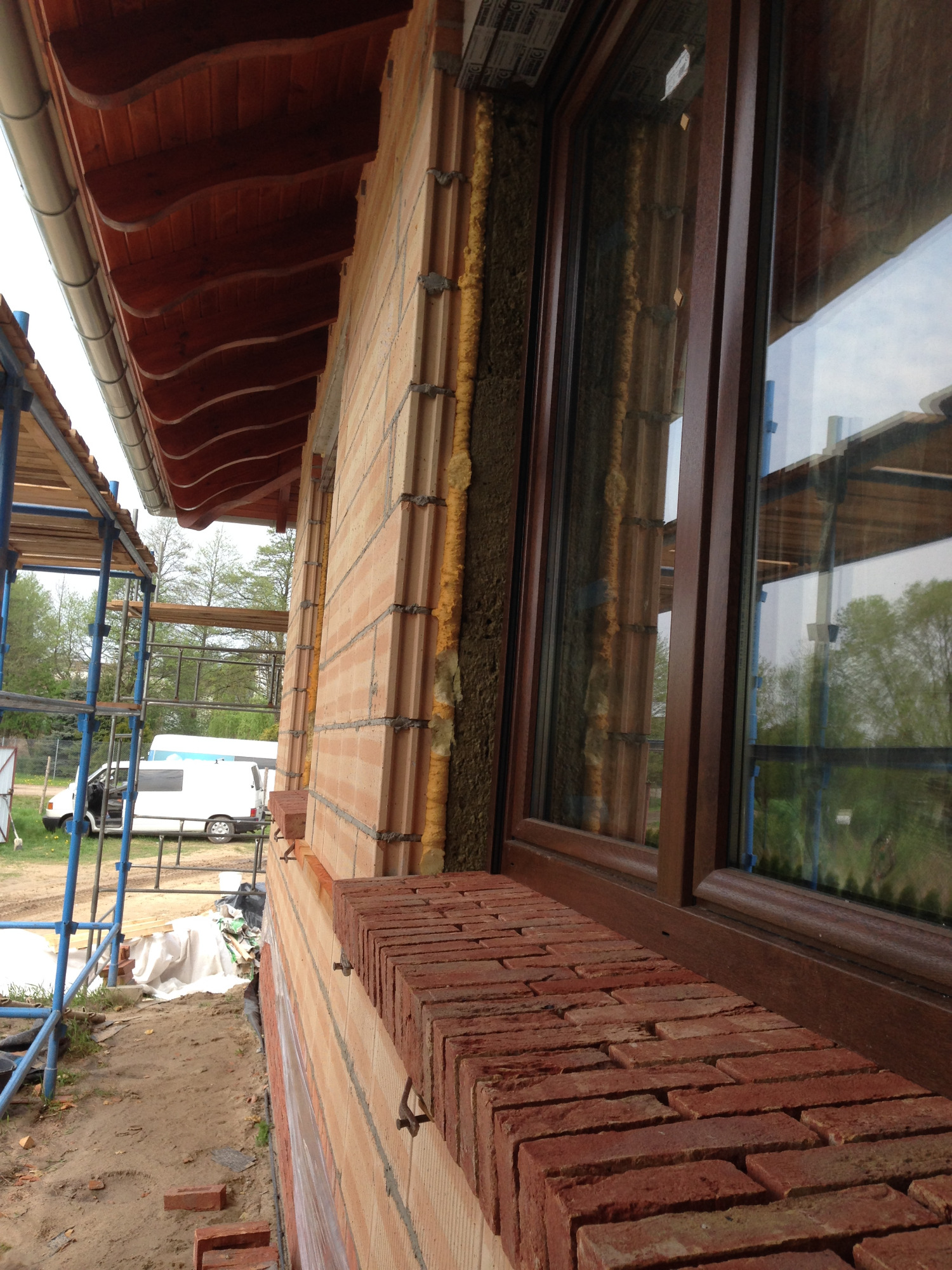 Fragment ściany z pustaków ceramicznych z zamontowanym oknem z brązowymi ramami, widoczna pianka izolacyjna i fragment drewnianej podbitki dachowej, na zewnątrz rusztowanie i zaparkowany biały bus.