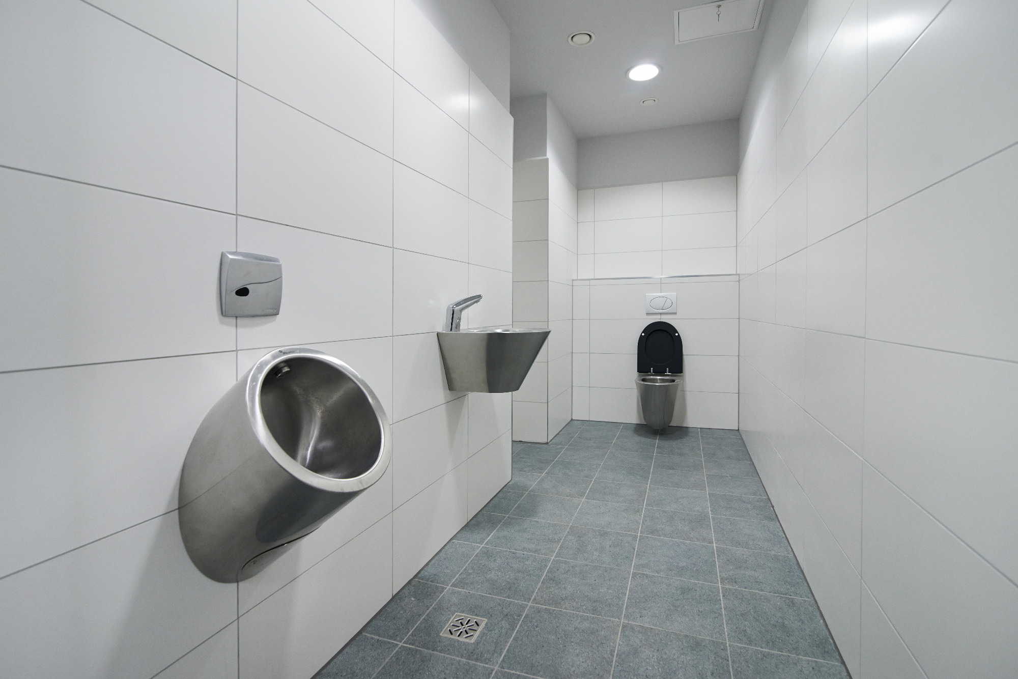 Wnętrze publicznej toalety z nowoczesnymi, metalowymi pisuarami i umywalką, wykończonej białymi płytkami i szarą podłogą.