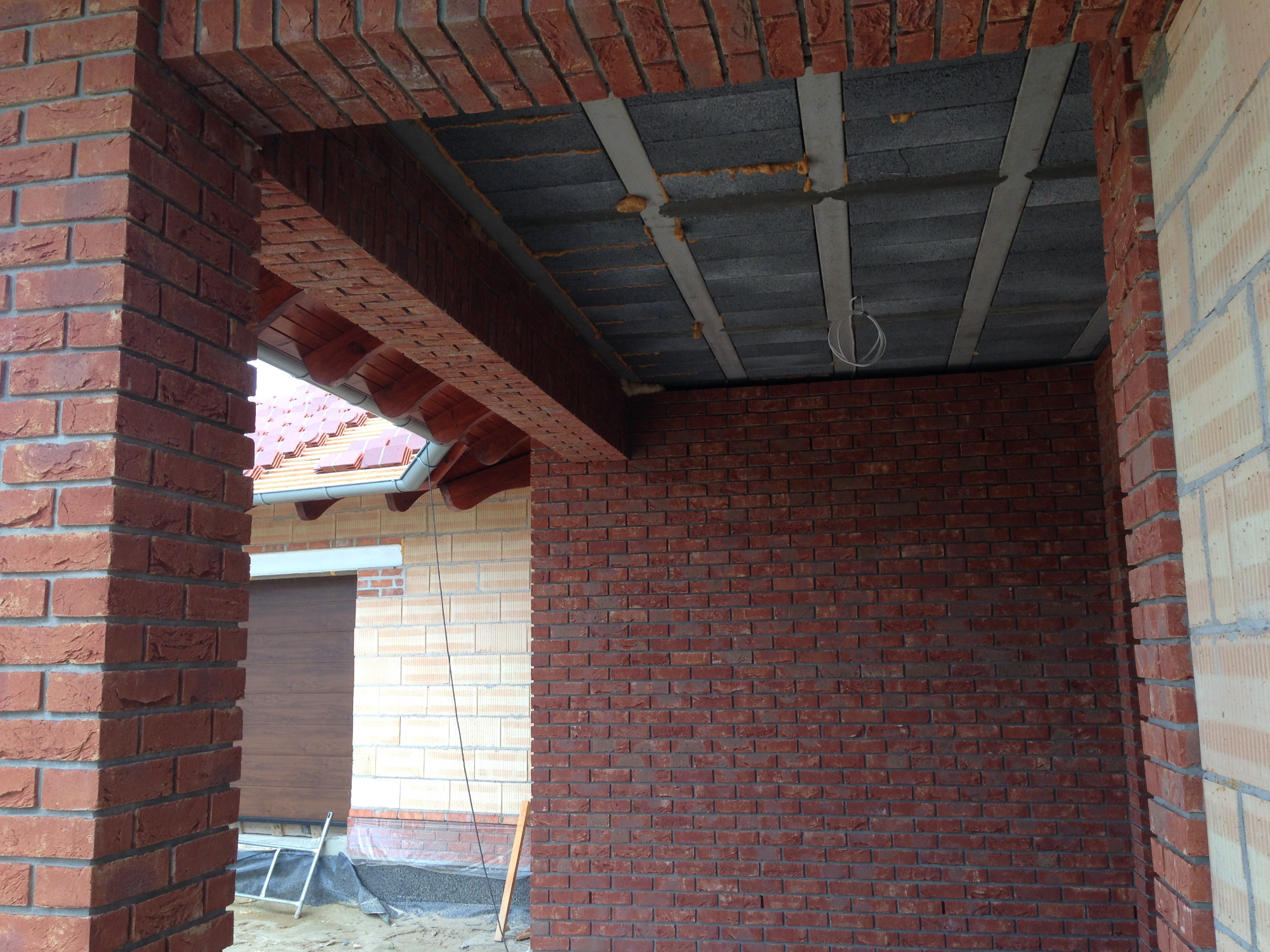 Surowa ściana z czerwonej cegły klinkierowej w trakcie budowy domu, widoczne nadproże z cegły i betonowy strop z instalacją elektryczną, fragment dachu i brama garażowa w tle.