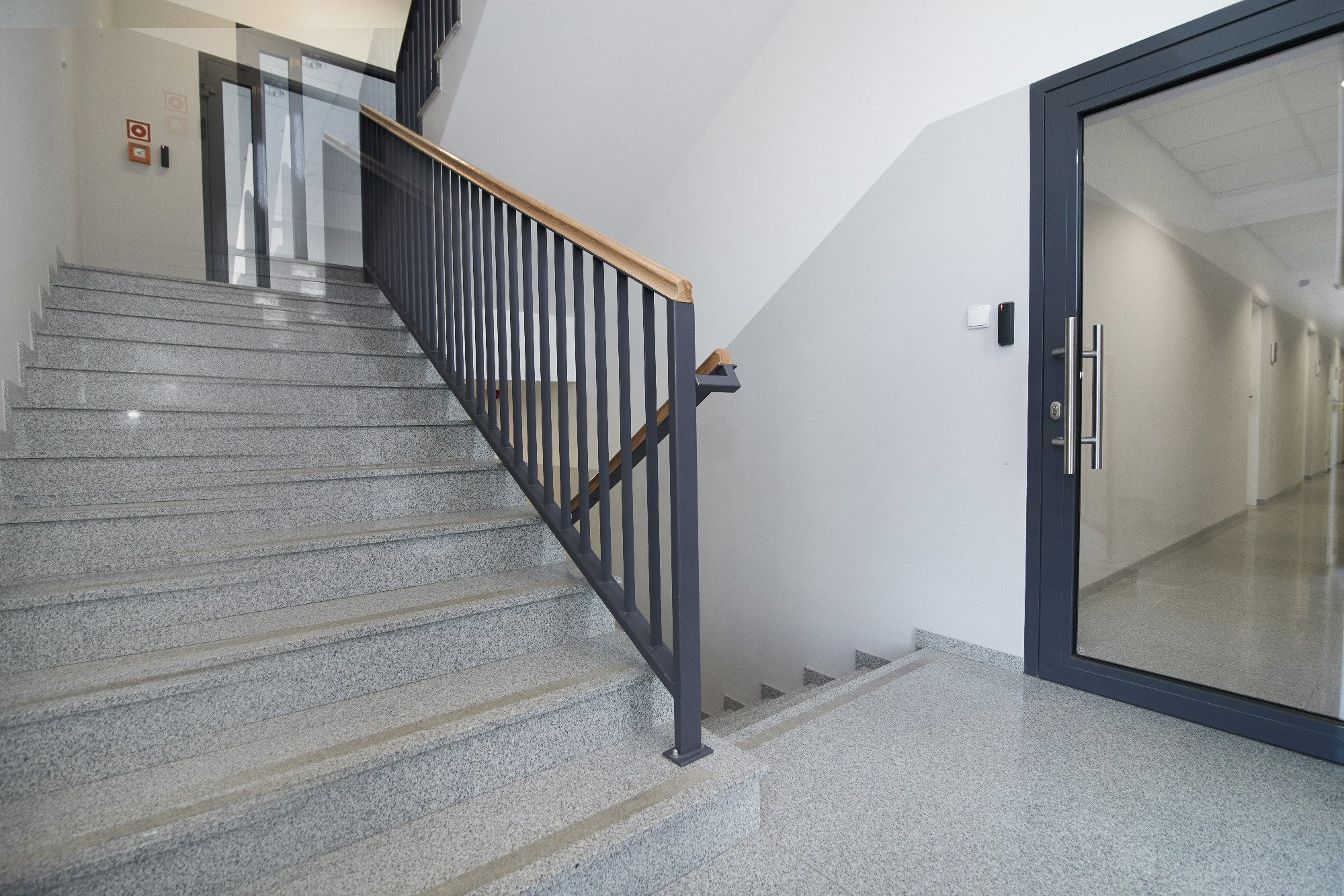 Schody z szarego lastryko z metalową balustradą i drewnianym poręczą w nowoczesnym budynku biurowym, widok na korytarz przez szklane drzwi.