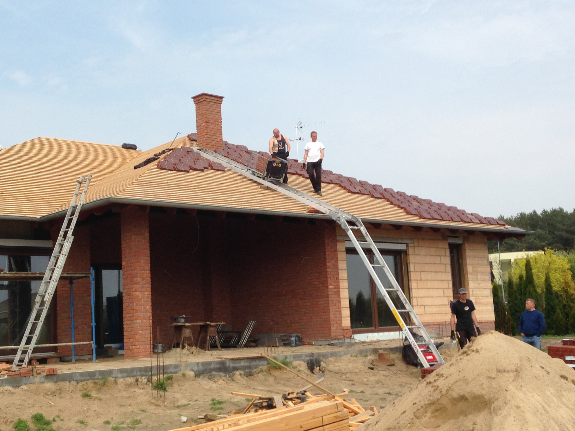 Montaż brązowej dachówki na nowo budowanym, jednopiętrowym domu z cegły; dwóch mężczyzn na dachu używa specjalnej windy pochyłej, trzeci stoi na ziemi obok sterty piasku.