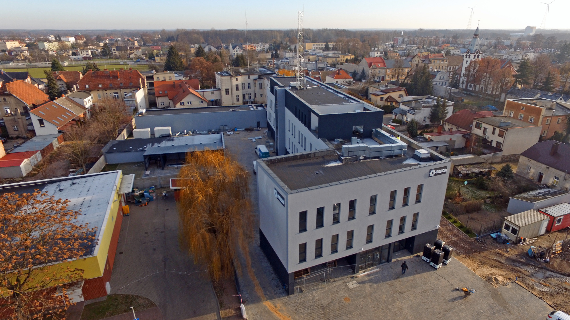 Widok z lotu ptaka na nowoczesny budynek komendy policji w Nowym Tomyślu, z szarą elewacją i czarnymi akcentami, otoczony brukowanym placem i elementami zagospodarowania terenu w trakcie budowy.