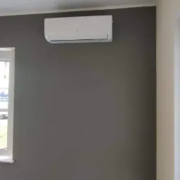 Biała klimatyzacja zamontowana na szarej ścianie w pokoju, widok z otwartego okna na samochód serwisowy na zewnątrz.