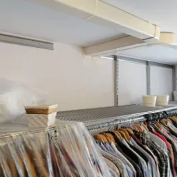 Wnętrze garderoby z zainstalowaną klimatyzacją kanałową pod sufitem, widoczny ruszt z ubraniami, doniczki i osłonięte folią przedmioty.