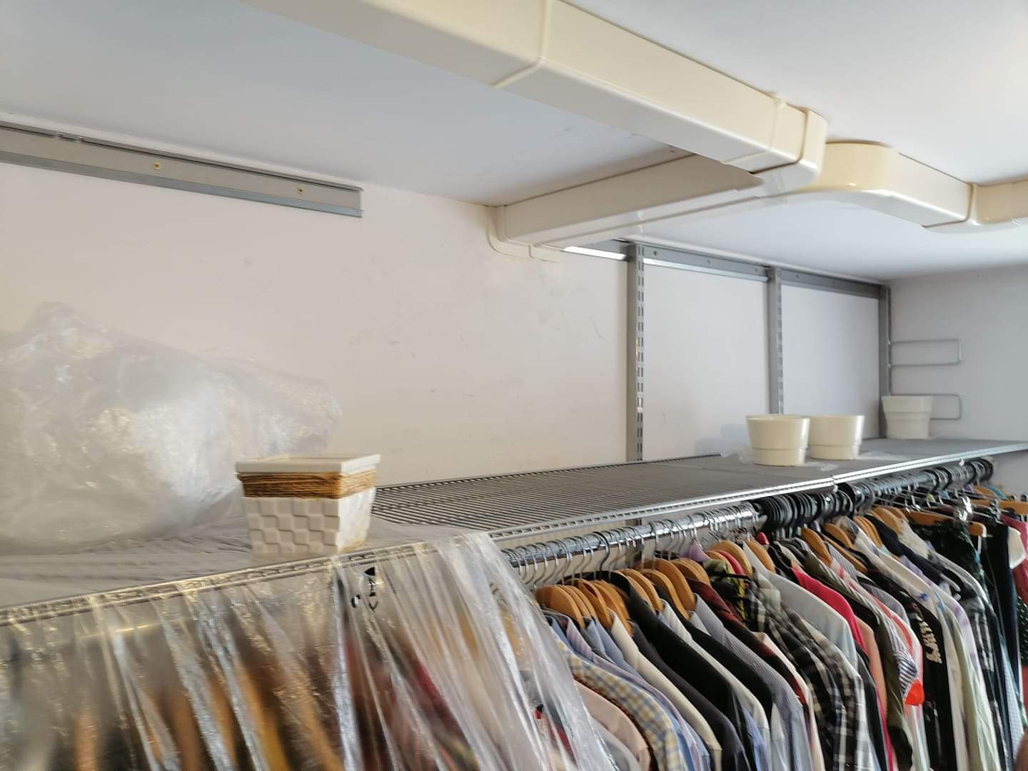 Wnętrze garderoby z zainstalowaną klimatyzacją kanałową pod sufitem, widoczny ruszt z ubraniami, doniczki i osłonięte folią przedmioty.