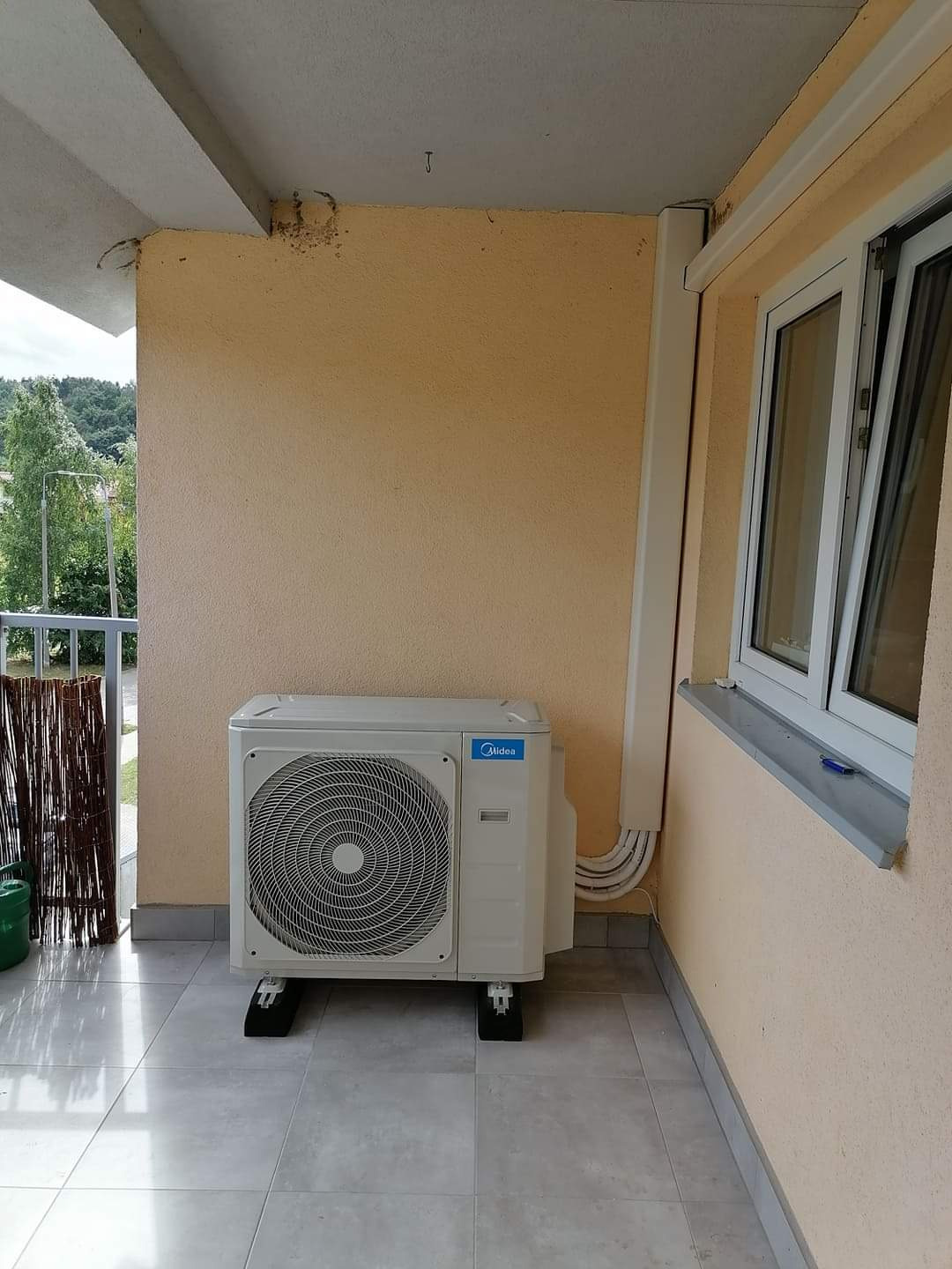 Zewnętrzna jednostka klimatyzacji Midea zamontowana na balkonie, widoczne przewody prowadzące do wnętrza budynku.