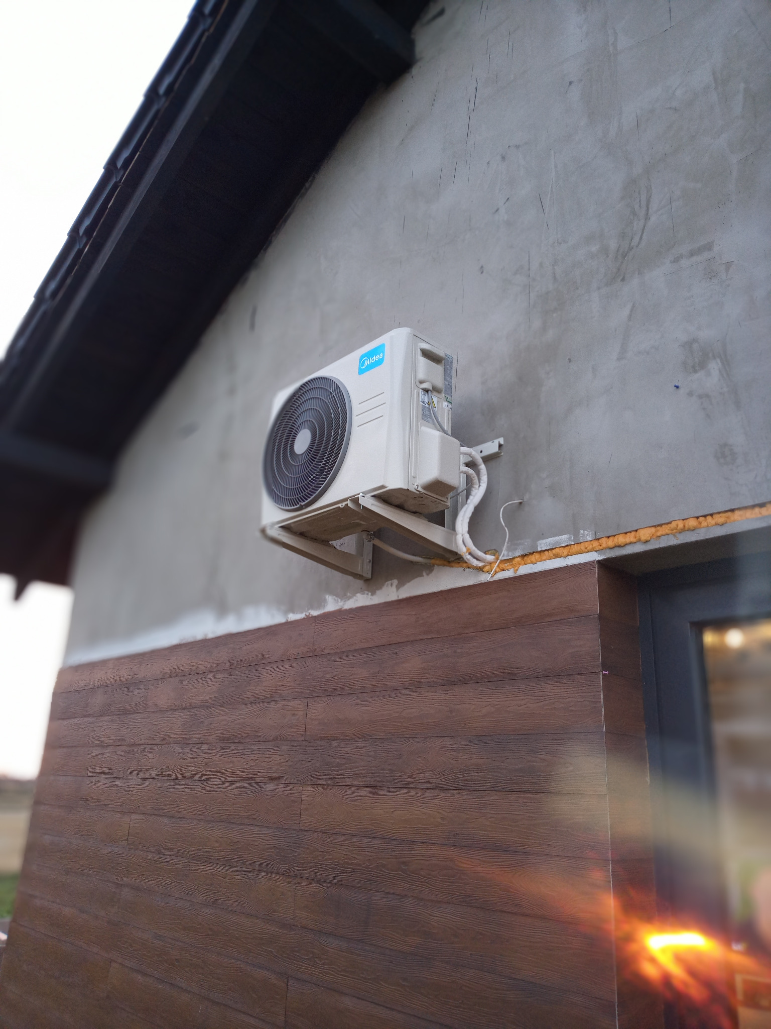 Zewnętrzna jednostka klimatyzatora Midea zamontowana na szarej ścianie domu z brązową okładziną, widoczne przewody i pianka montażowa wokół okna.
