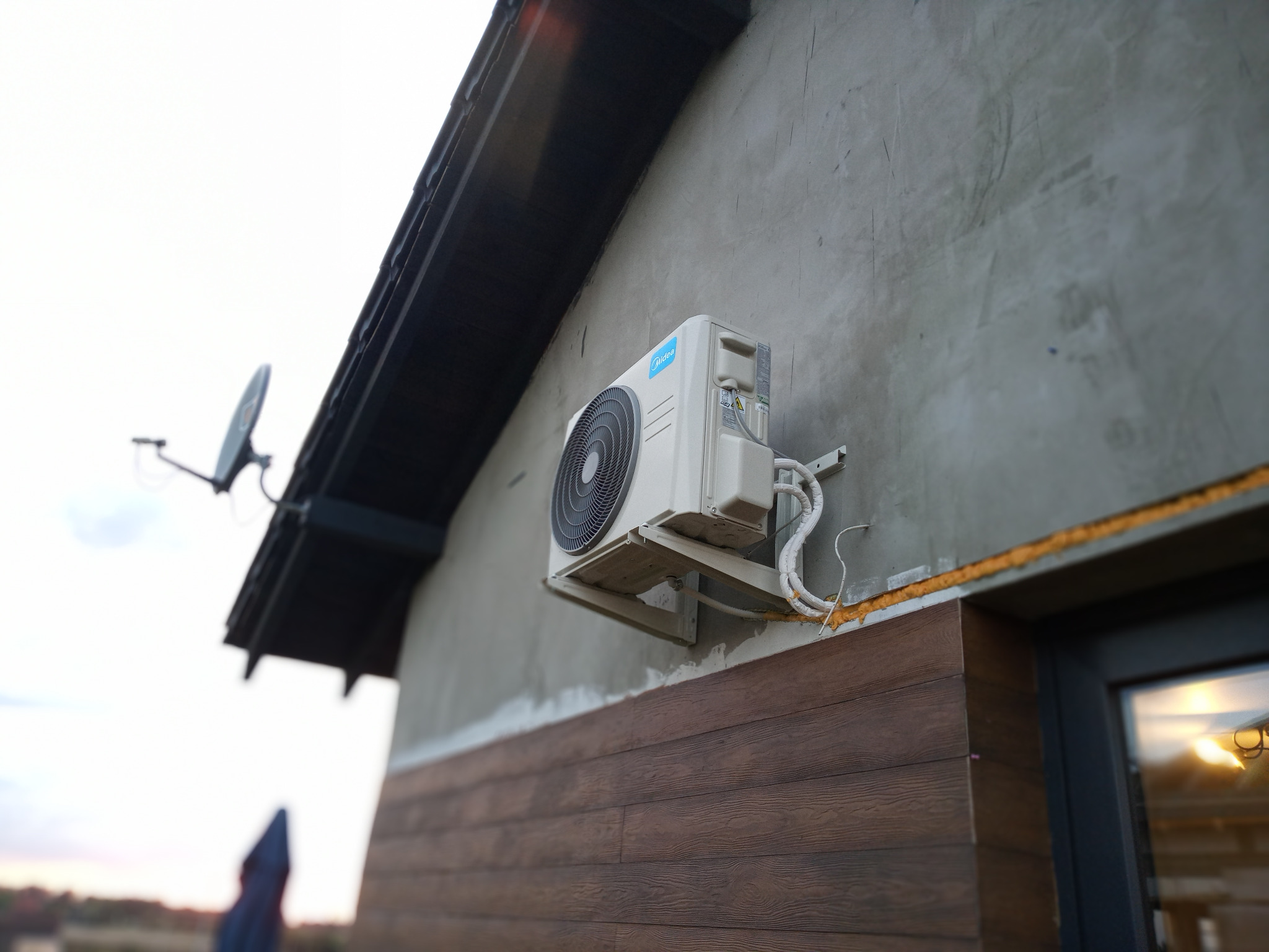 Zewnętrzna jednostka klimatyzacji Midea zamontowana na elewacji domu z drewnianą okładziną, widoczna antena satelitarna w tle.
