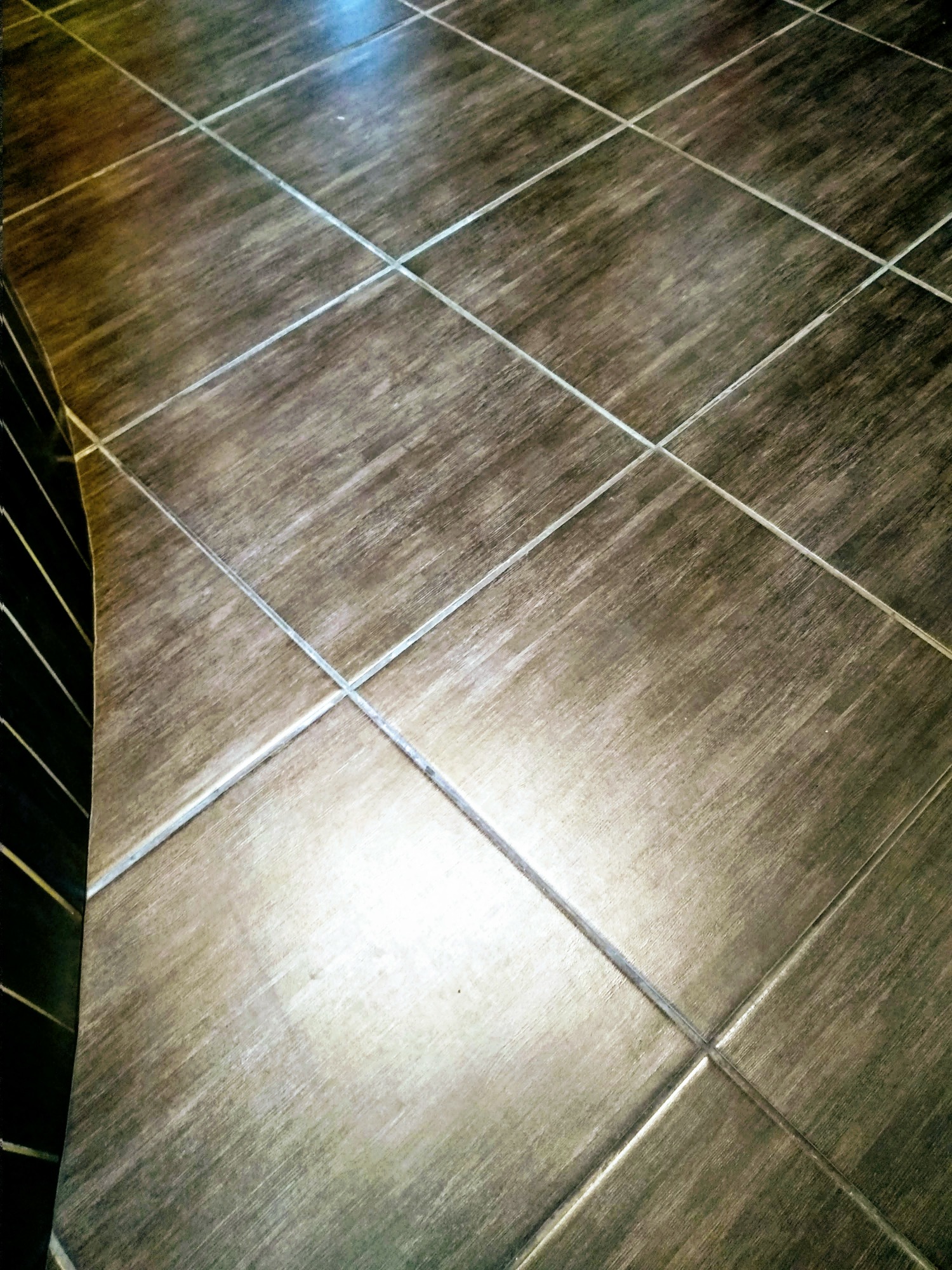 Ułożone pod kątem brązowe płytki podłogowe z jasnoszarą fugą, widoczne fragmenty czarnej ściany z pionowymi paskami płytek po lewej stronie.