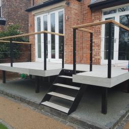 SK Gardens - Nowoczesny taras z szarych desek kompozytowych, czarnych metalowych słupków i jasnych drewnianych poręczy, z niskimi schodami prowadzącymi na poziom gruntu, na tle ceglanego domu z białymi drzwiami...
