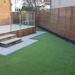 SK Gardens - Zewnętrzny taras z szarych płyt, schodami, przeszkloną balustradą i drewnianym ażurowym ogrodzeniem na sztucznej trawie.