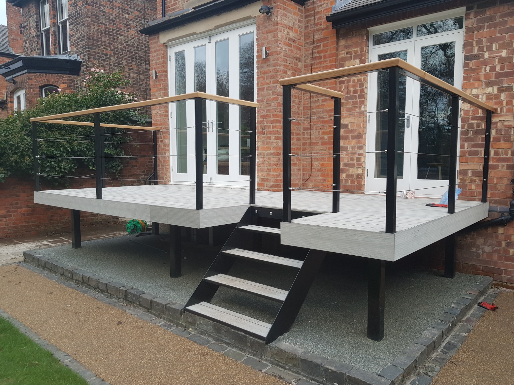 Nowoczesny taras z szarych desek kompozytowych, czarnych metalowych słupków i jasnych drewnianych poręczy, z niskimi schodami prowadzącymi na poziom gruntu, na tle ceglanego domu z białymi drzwiami...