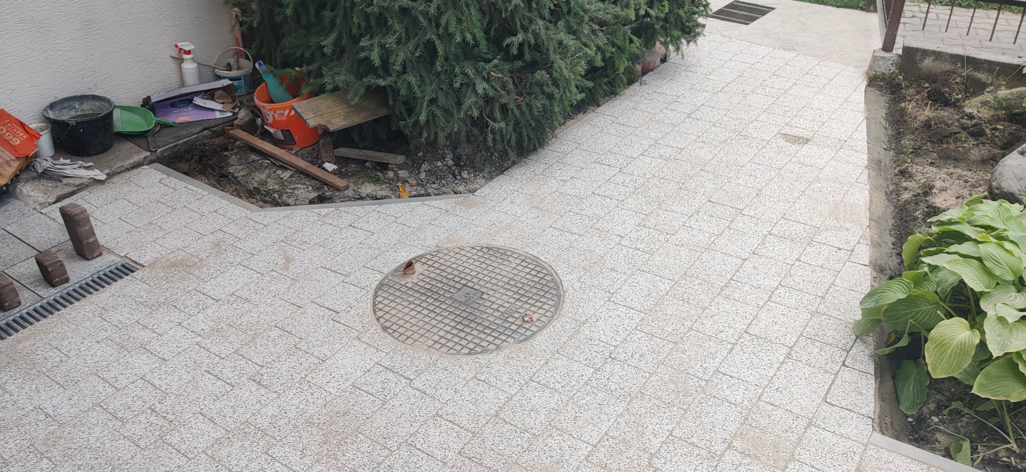 Ułożona jasna kostka brukowa na posesji, widoczny właz kanalizacyjny, krawężnik, rabata z roślinami i narzędzia budowlane w tle.