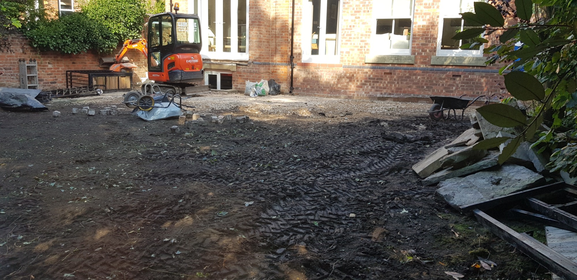 Przygotowanie terenu pod budowę: mini koparka, taczka, kostka brukowa i kamienie na błotnistym podłożu przed ceglanym budynkiem z białymi oknami.