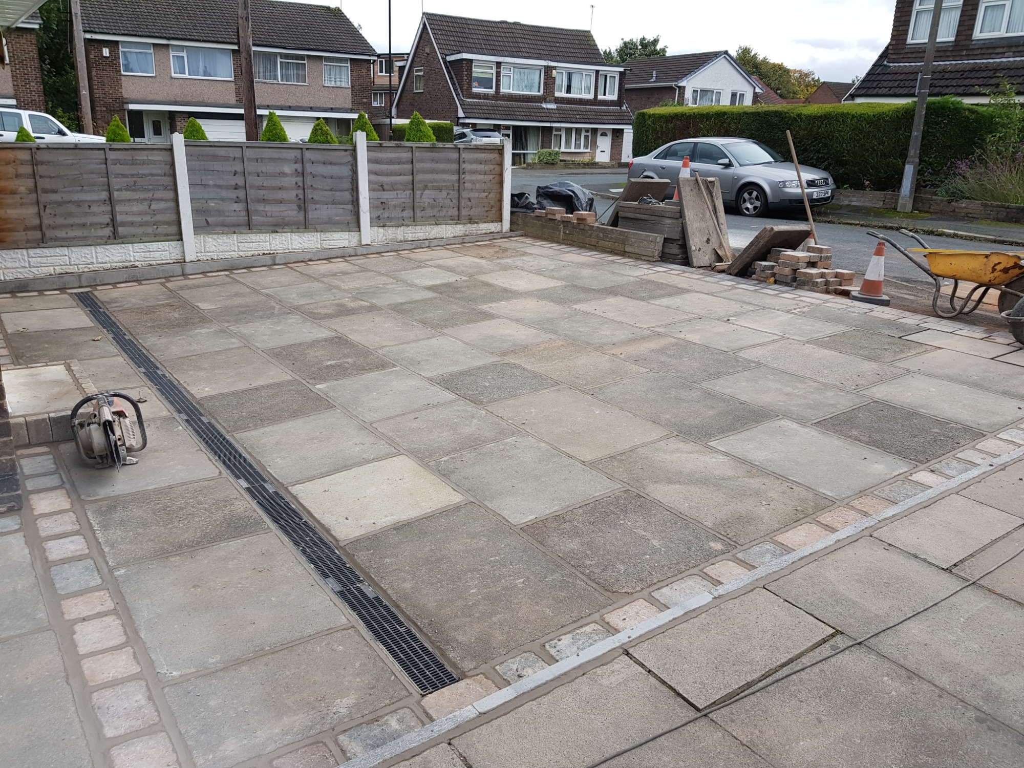 Ułożona nawierzchnia z szarych płyt betonowych z obrzeżami z cegły i odpływem liniowym, widoczne narzędzie do cięcia i taczka, domy w tle.