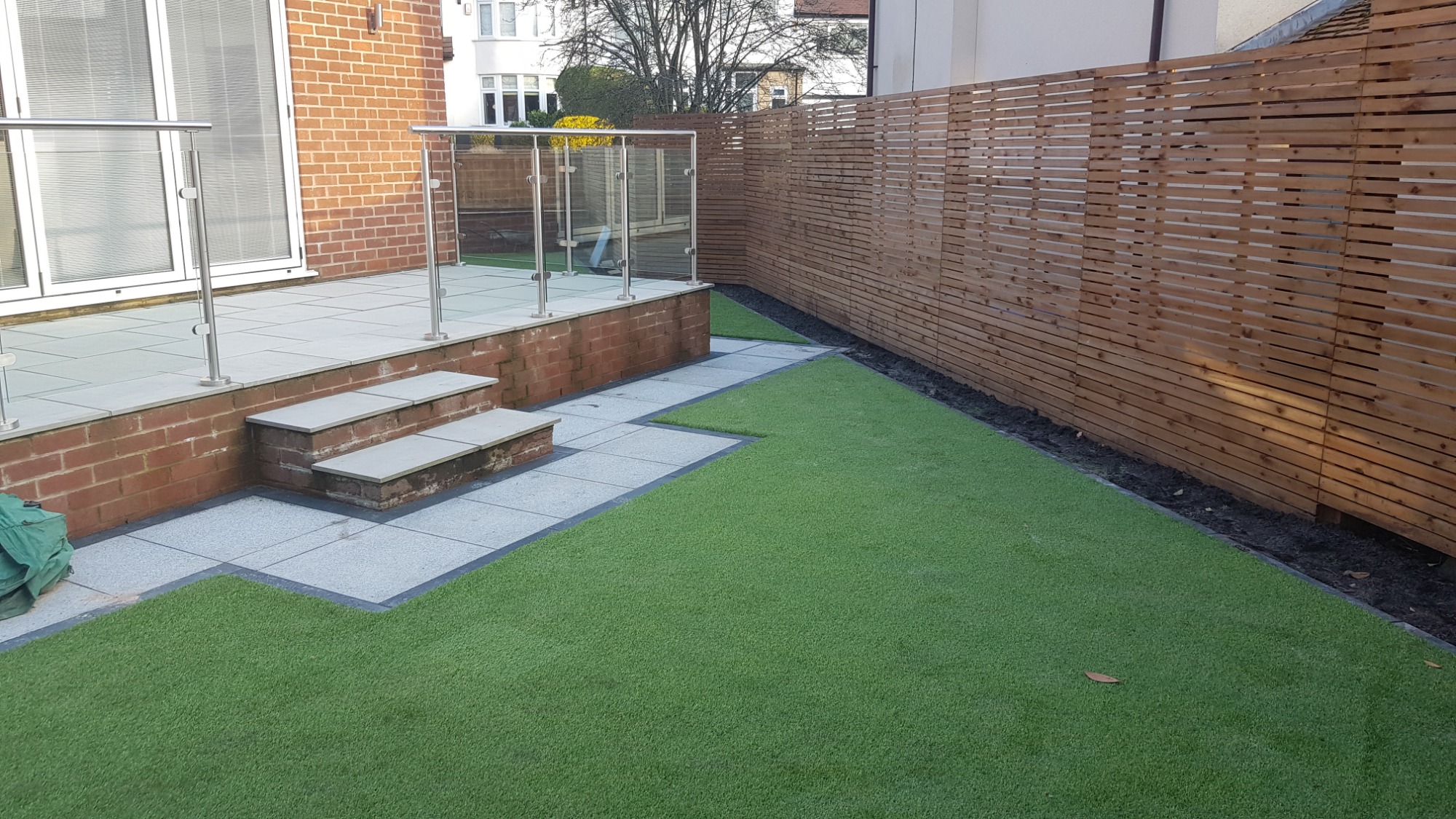 Zewnętrzny taras z szarych płyt, schodami, przeszkloną balustradą i drewnianym ażurowym ogrodzeniem na sztucznej trawie.