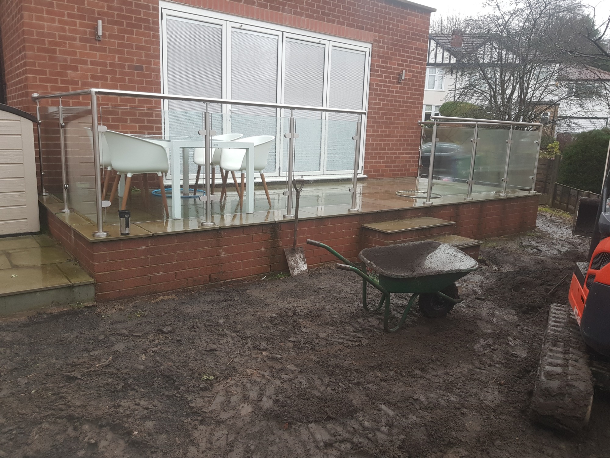 Wykończony taras z kostki brukowej z szklaną balustradą, widoczne białe krzesła ogrodowe i stół, na pierwszym planie błotniste podłoże z taczką i łopatą oraz częściowo widoczna koparka.