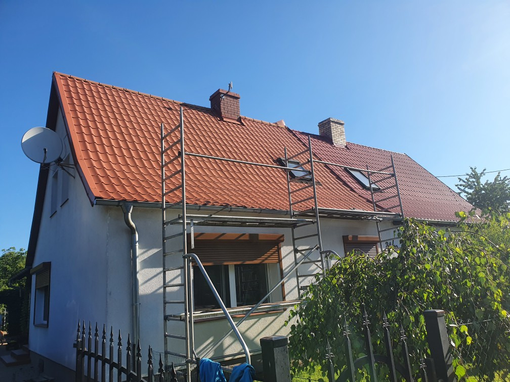 Dom jednorodzinny z nowym, czerwonym dachem z dachówki ceramicznej, otoczony rusztowaniem i zielenią, widok z dołu w słoneczny dzień.