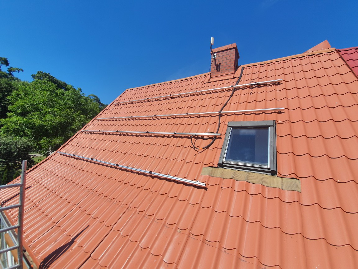 Czerwony dach z dachówki z zamontowanymi aluminiowymi szynami montażowymi pod panele słoneczne, widoczne okno dachowe i komin z anteną, błękitne niebo w tle.