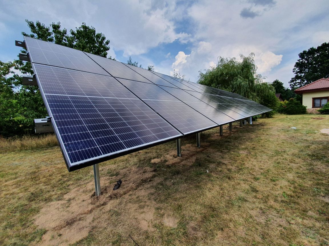 Rząd paneli słonecznych ustawionych na metalowych wspornikach na trawiastym terenie, z domem w tle i pochmurnym niebem.