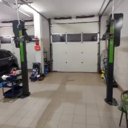 Ciemny SUV na podnośniku w warsztacie samochodowym, widoczne dwa podnośniki kolumnowe, w tle brama garażowa i regał z narzędziami.