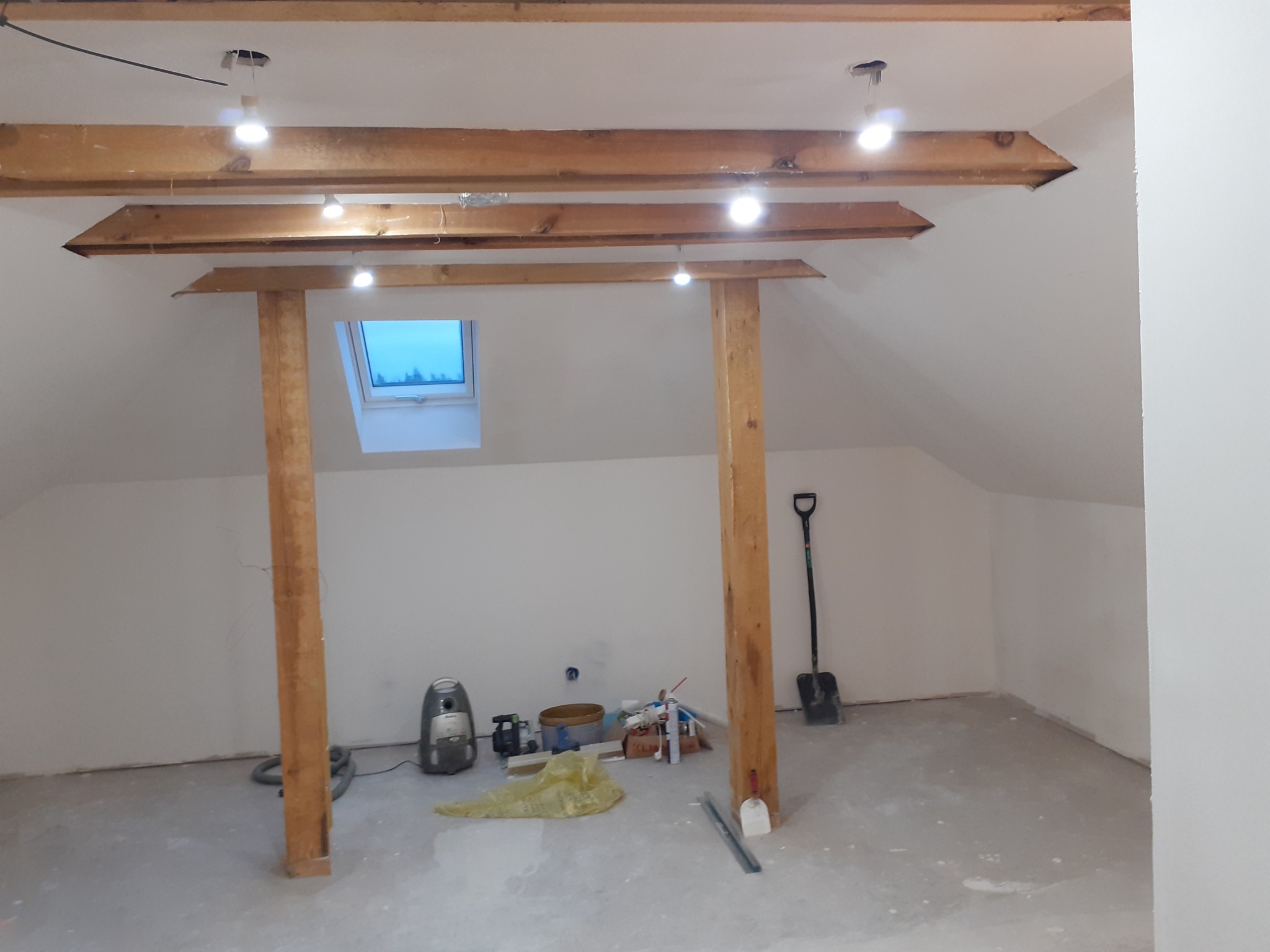 Poddasze w trakcie prac wykończeniowych; widoczne drewniane belki stropowe z oświetleniem, okno dachowe, przygotowane ściany do malowania, odkurzacz, szpachla i łopata.