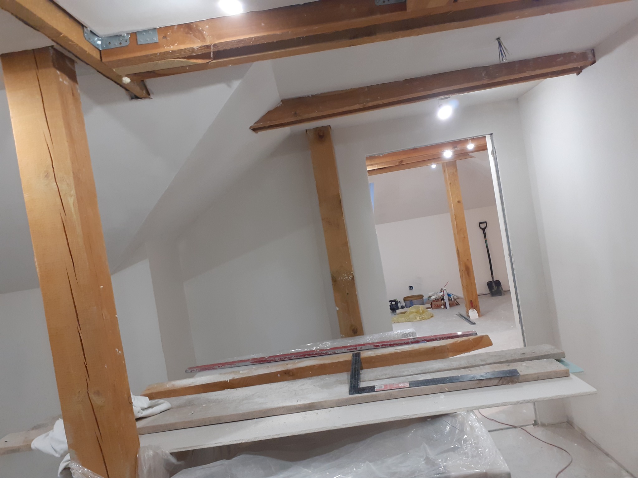 Wnętrze poddasza w trakcie prac wykończeniowych: drewniane belki konstrukcyjne, pionowe słupy, przygotowane płyty gipsowe z narzędziami na tymczasowym podeście, widok na kolejne pomieszczenie...